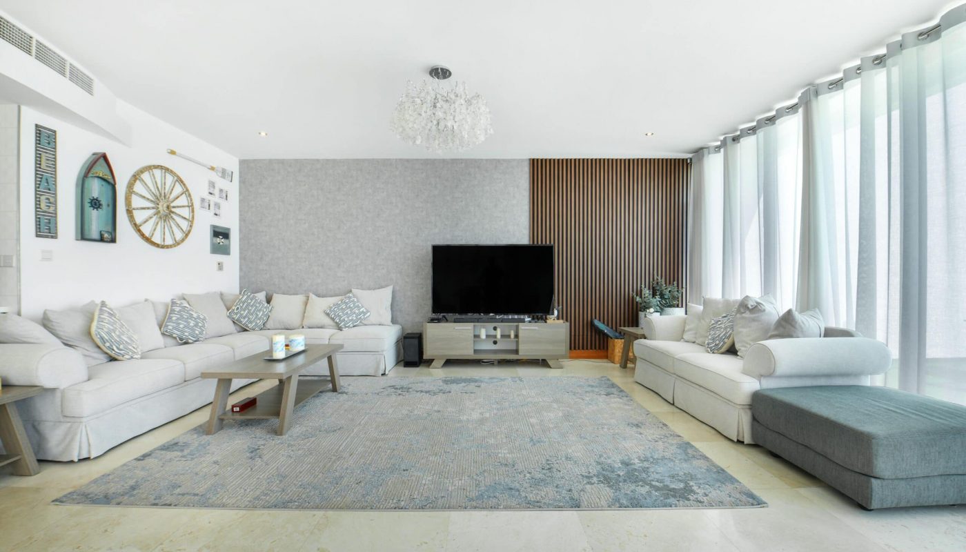 Spacious living room with two sofas, a flat-screen TV, a grey-blue rug, and large windows with sheer curtains. Walls are decorated with art and a ship's wheel.
