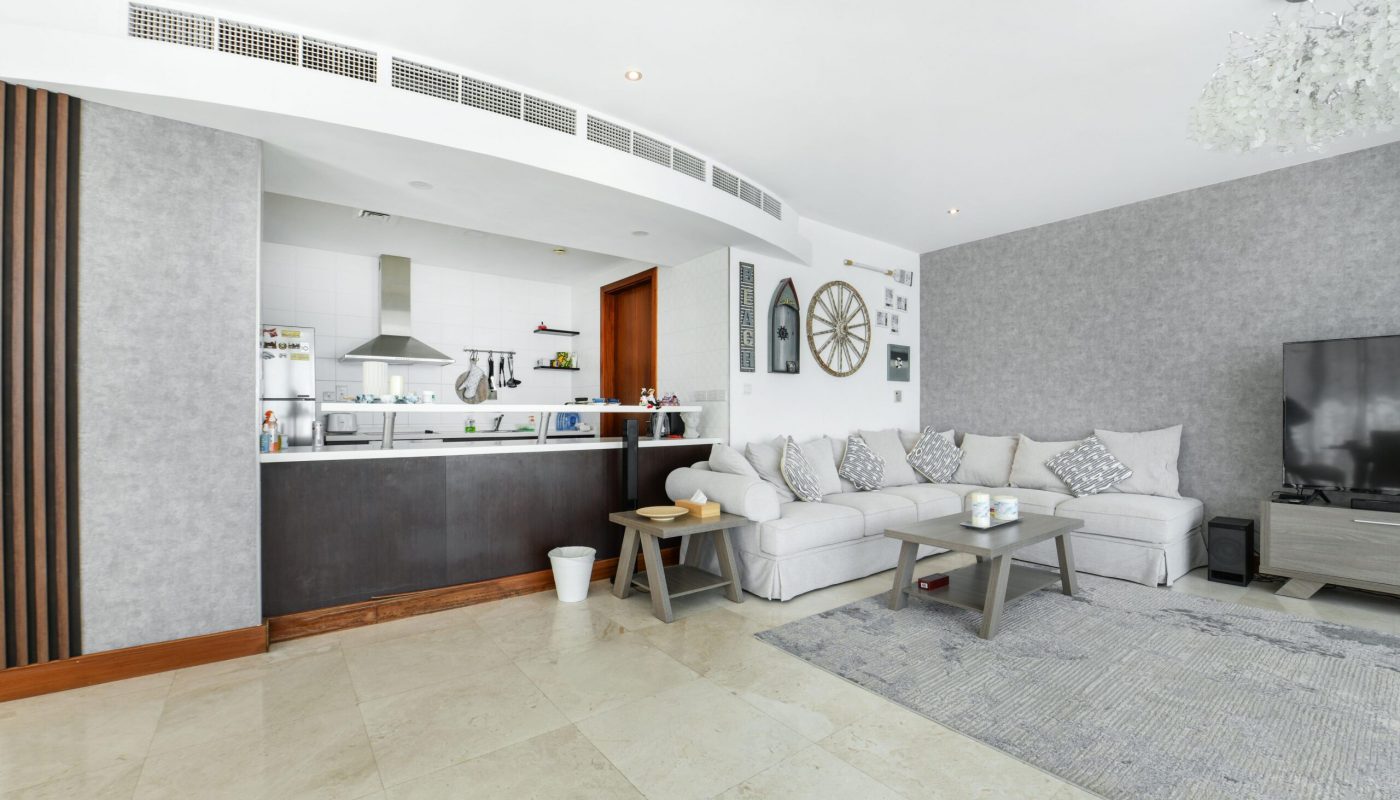 Modern open-plan living room and kitchen with white sectional sofa, gray rug, large TV, kitchen counter with stools, wall decor, and tiled floor.