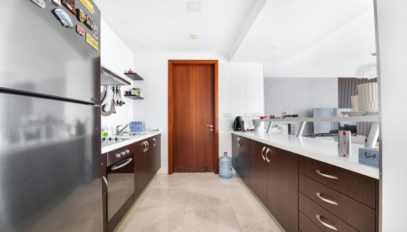 Modern kitchen with stainless steel fridge, dark wood cabinets, white countertops, and beige tile floor. A wooden door at the end, with a wall mirror reflecting the opposite side.