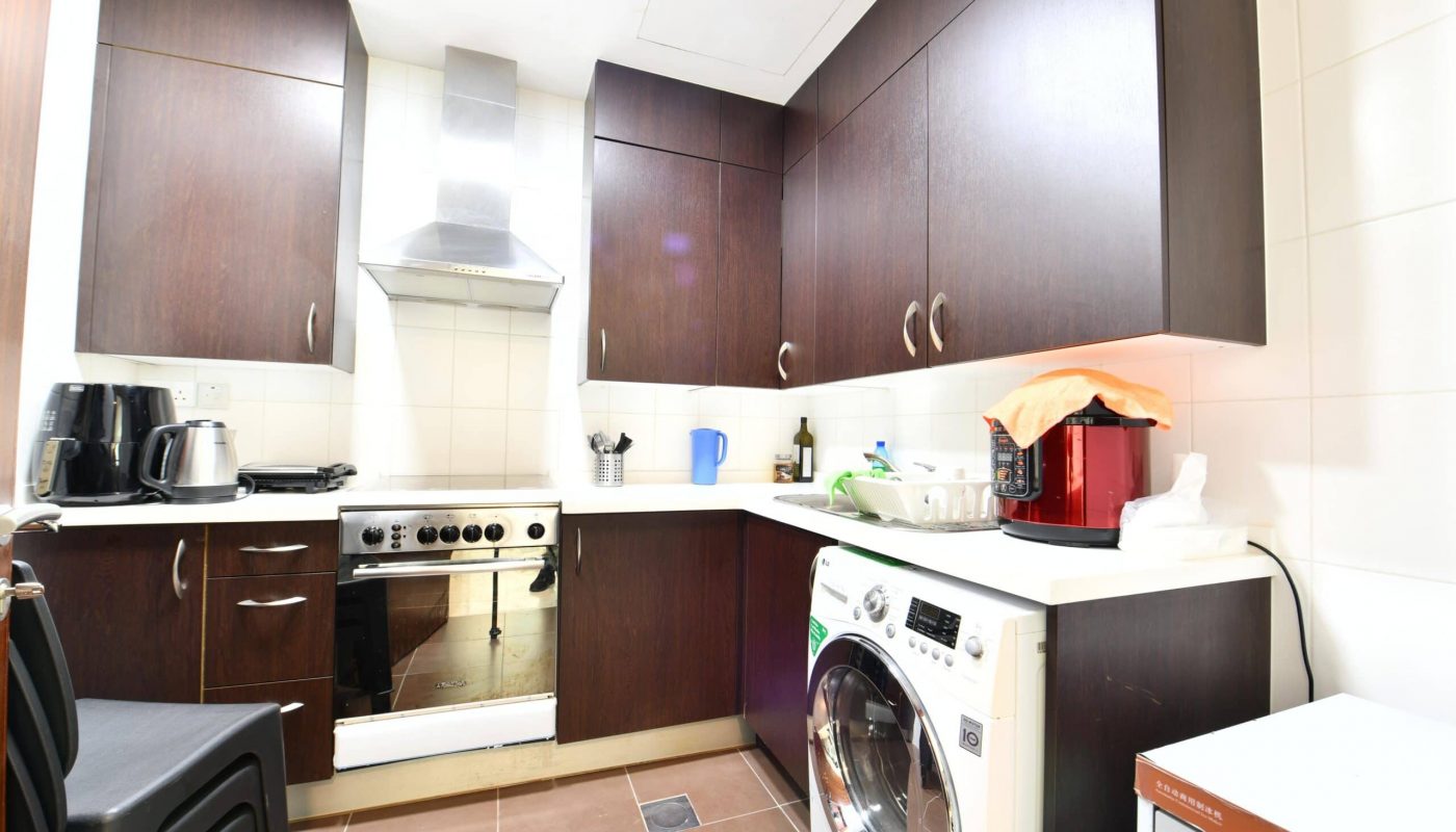 Compact kitchen with dark wooden cabinets, a stainless steel oven, washing machine, and small appliances on the counter. White tiled backsplash and brown floor tiles.