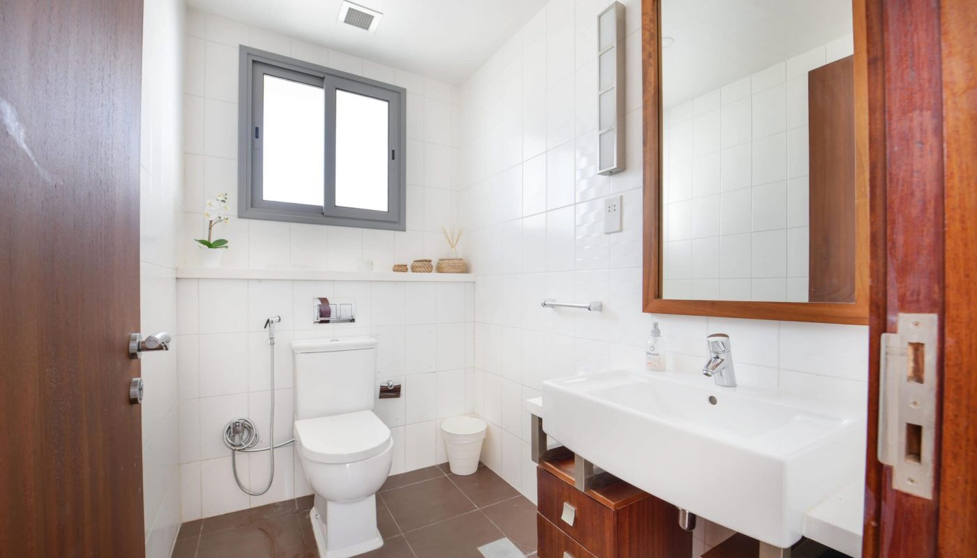Modern bathroom with a white toilet, white sink, and wooden cabinets. Brown tiled floor, white walls, large mirror, and a window above the toilet. Accessories on shelf and sink.