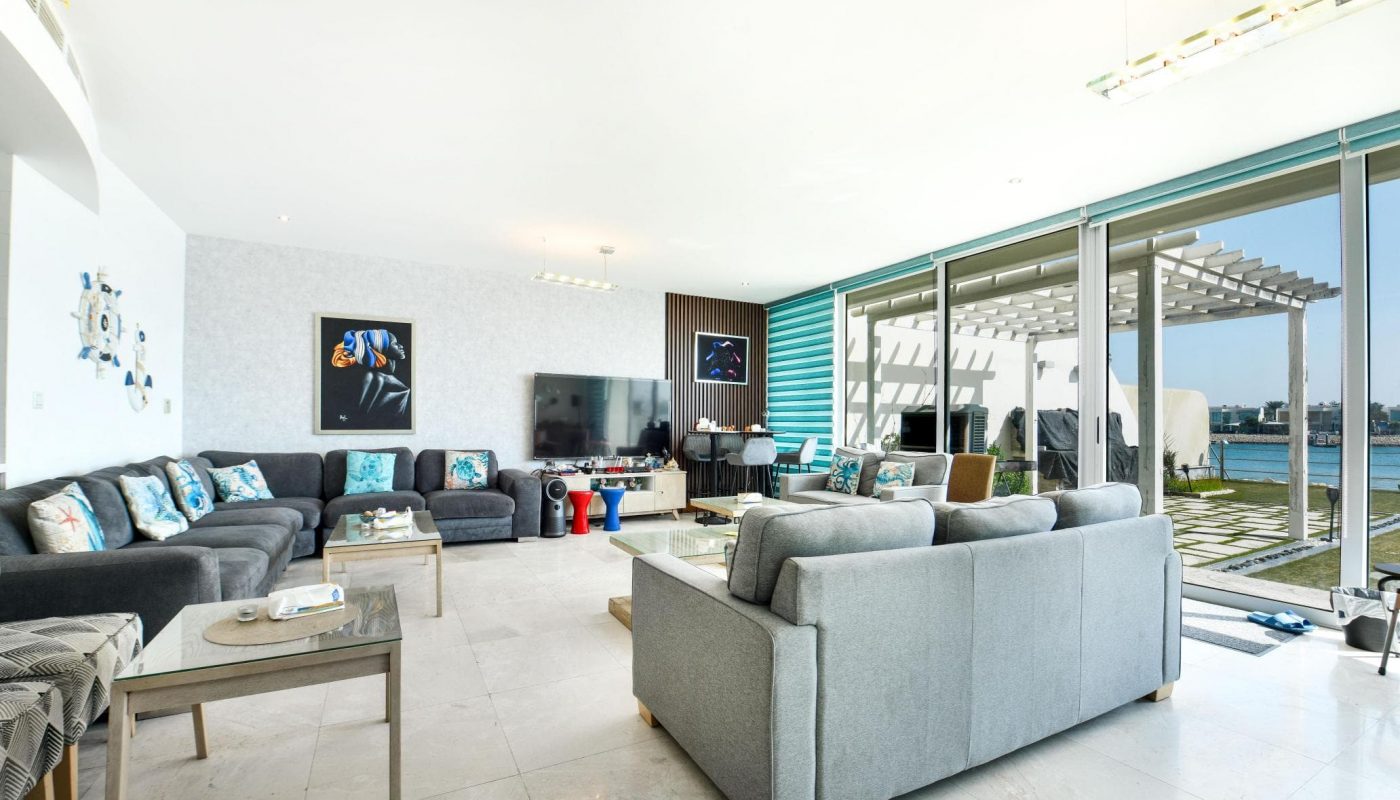 Spacious, modern living room with gray sofas, a large TV, and glass doors leading to a patio overlooking the water. Bright with natural light and contemporary decor.