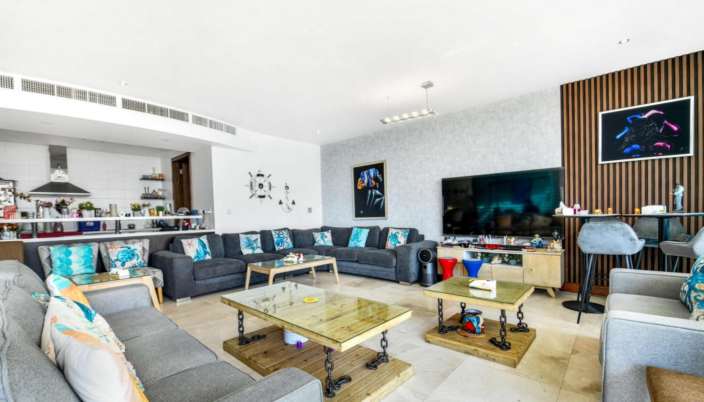 Spacious living room with multiple gray sofas, a large TV, and a glass coffee table. Art decorates the walls. An open kitchen is visible in the background.