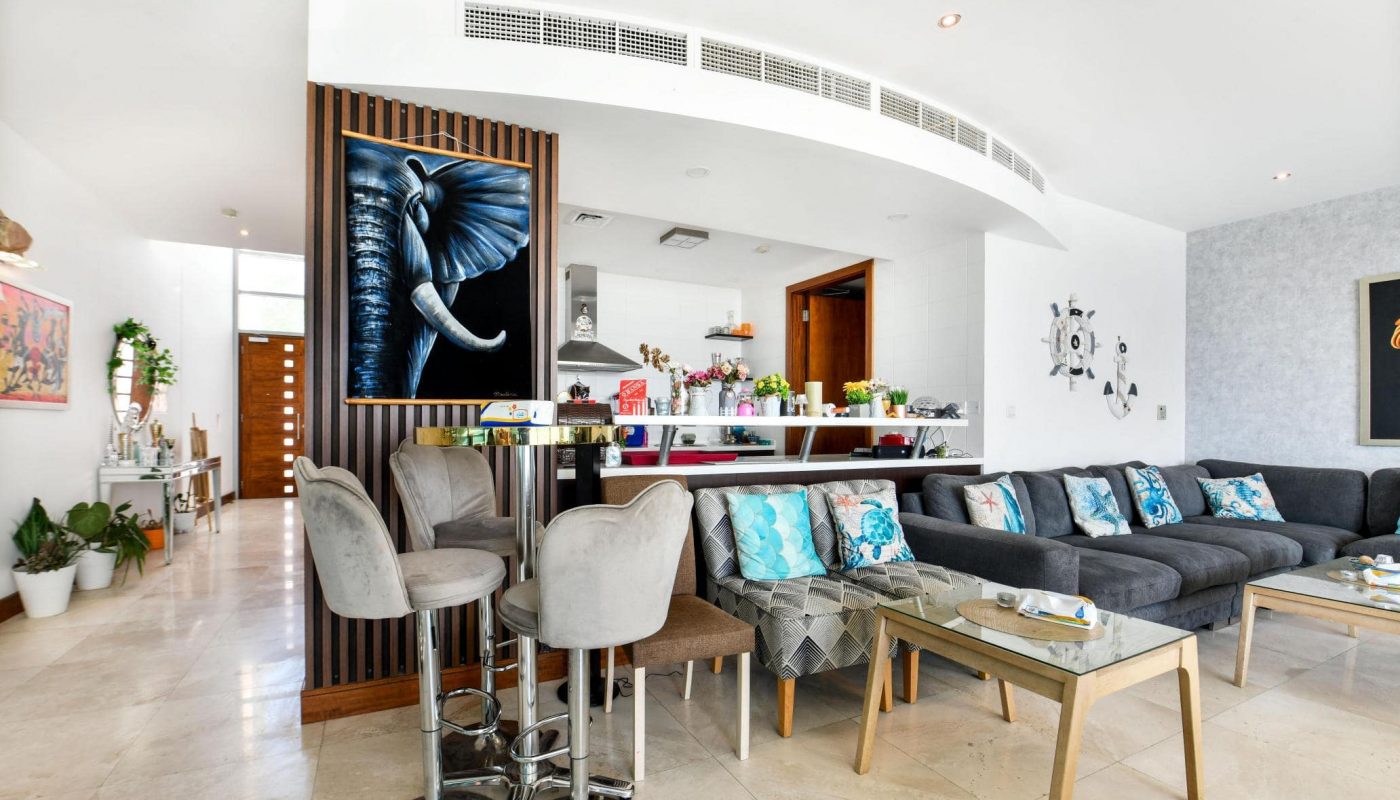 Modern living room with a gray sectional sofa, patterned cushions, and an elephant painting. A kitchen area with bar stools is visible, alongside indoor plants and a light marble floor.
.