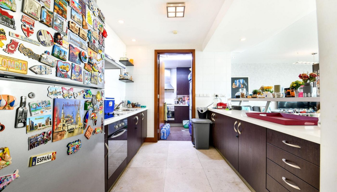 Modern kitchen with dark wood cabinets, stainless steel fridge covered in magnets, light countertops, and a tiled floor. A doorway leads to another room at the back.