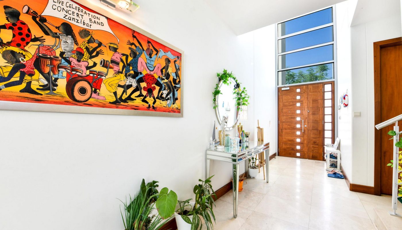 Bright hallway with large colorful painting on the left wall, a console table with a round mirror, and several potted plants. Modern wooden door with horizontal glass panels in the background.