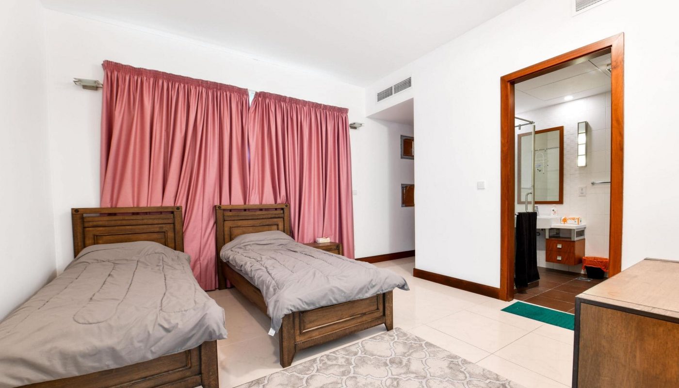 A bedroom with two wooden single beds, gray bedding, pink curtains, a dresser, and an open door leading to a bathroom with a sink and shower.