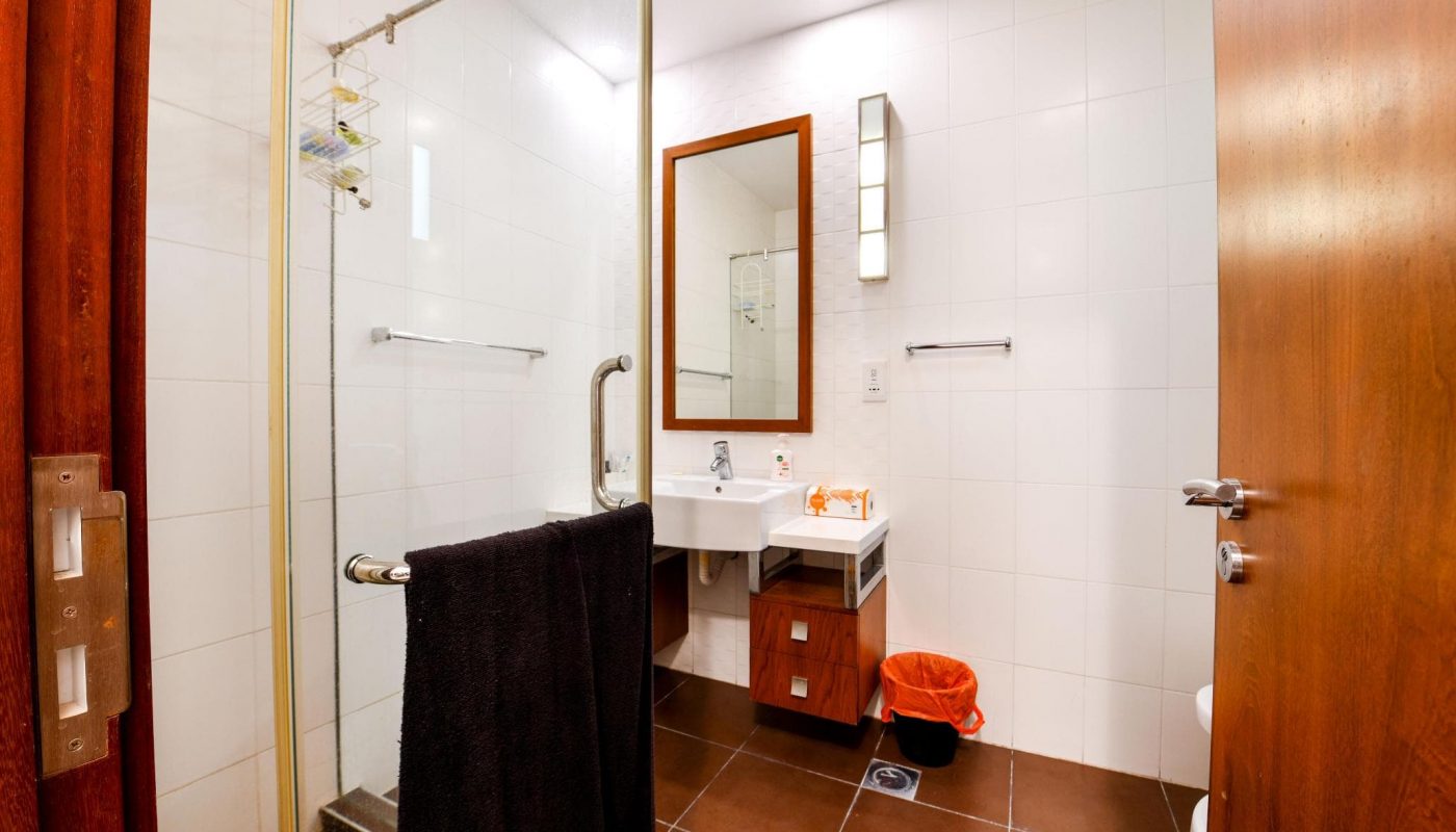 Modern bathroom with glass shower, white tiles, wood accents, a sink with a mirror, towel rack, black towel, and orange trash bin on a brown tiled floor.