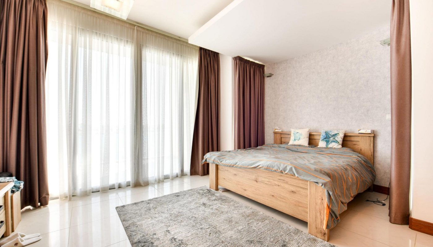A bedroom with a large wooden bed, gray and blue bedding, windows with sheer and brown curtains, and a gray rug on a glossy floor.