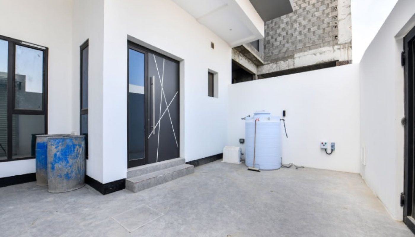 A white outdoor area with a modern glass door, a large water tank, and a blue industrial barrel on a concrete surface.