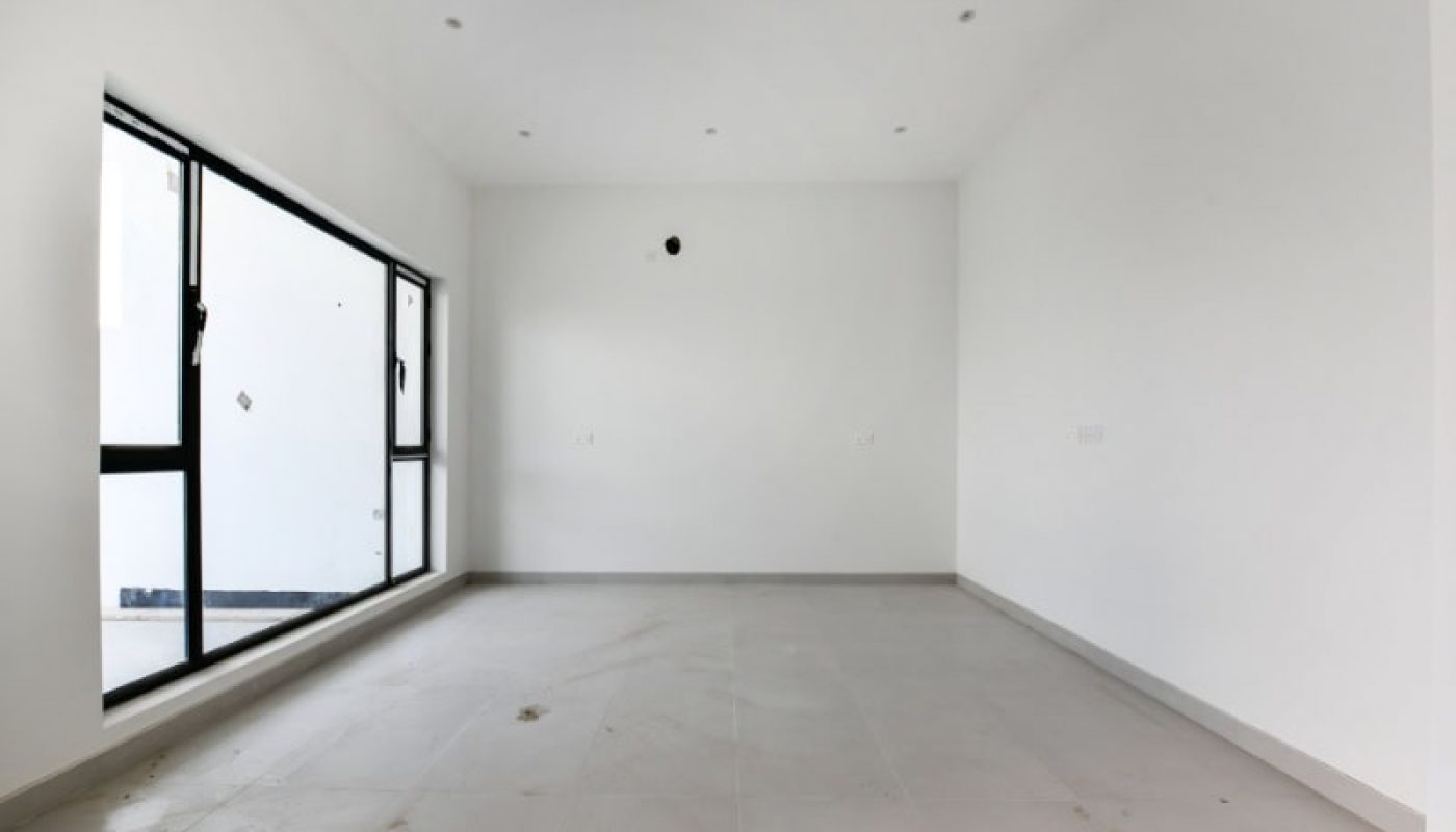 Empty room with white walls, large window on the left, and recessed ceiling lights. The floor is light-colored and slightly dusty.