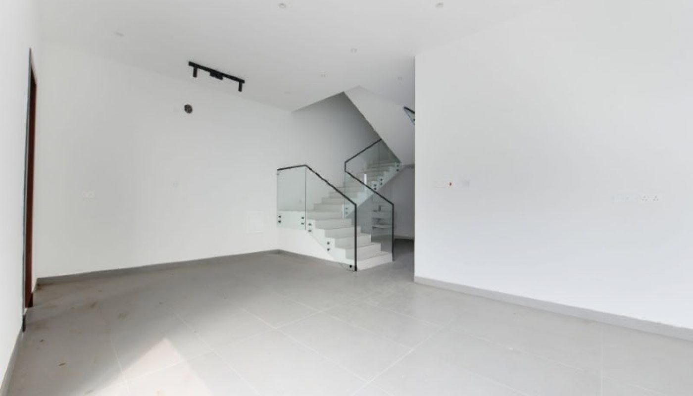 Empty room with white walls and tiled floor, featuring a staircase with glass railing leading to an upper level. Ceiling lights are installed, and a single door is partially open.