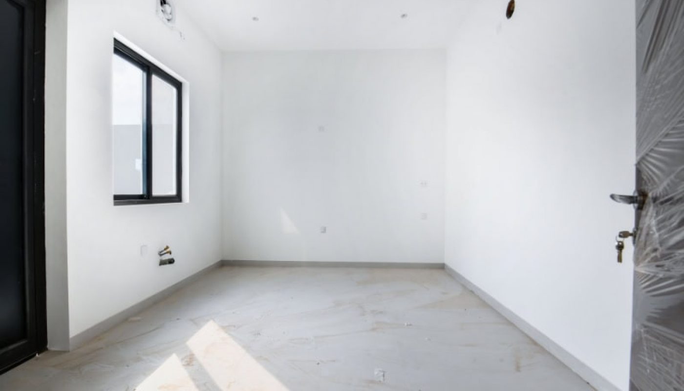 Empty, newly constructed white room with a tiled floor, a window on the left wall, and a black door on the right side. Dust and plaster remnants are scattered on the floor.