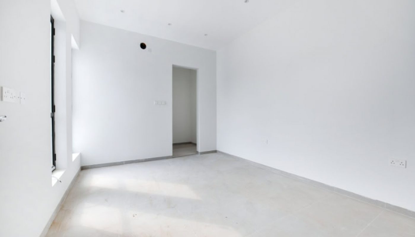 An empty, bright room with white walls and a tiled floor. A door leads to another space, and natural light enters through a window on the left.