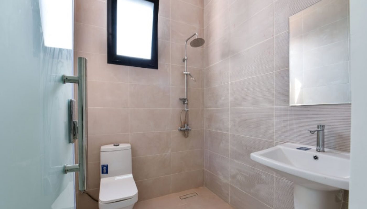 Minimalist bathroom with beige tiles, featuring a toilet, wall-mounted shower, rectangular mirror, and a sink with a silver faucet. A frosted glass door and a small window provide light.