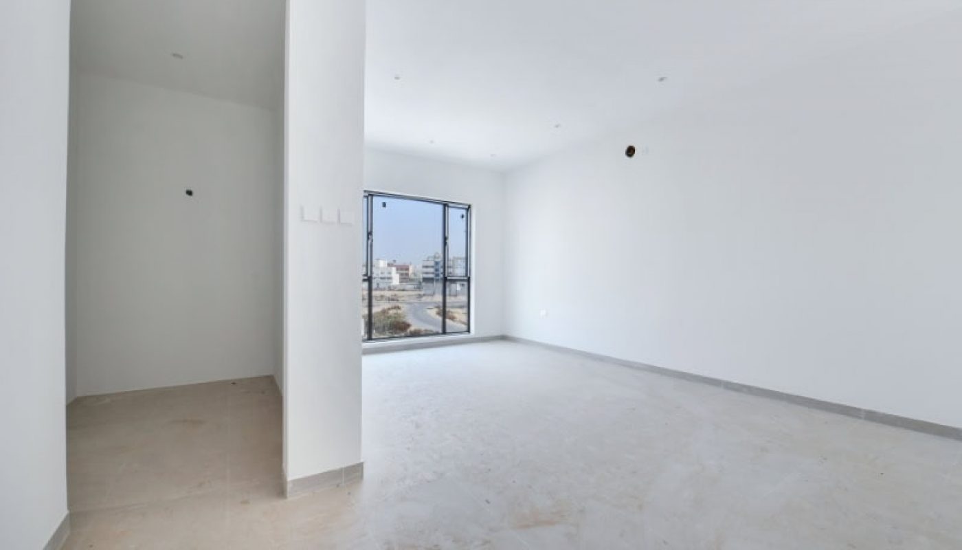 Empty white room with a large window, beige tiled floor, and recessed ceiling lights. An adjacent small area to the left has bare walls and an uninstalled fixture.