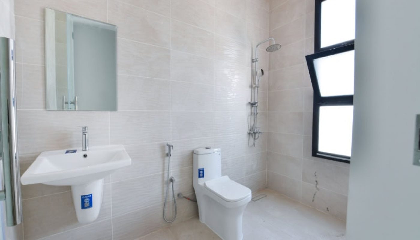 Bathroom with a wall-mounted sink, a toilet, and a shower area. The walls and floor are tiled in a light color. The room has a large window on the right side.