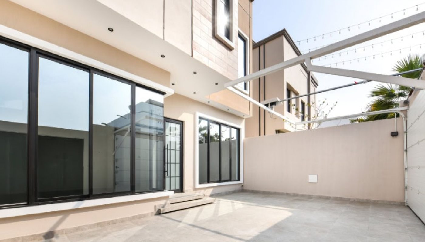Modern building exterior with large windows, beige walls, and spacious tiled patio under clear sky.