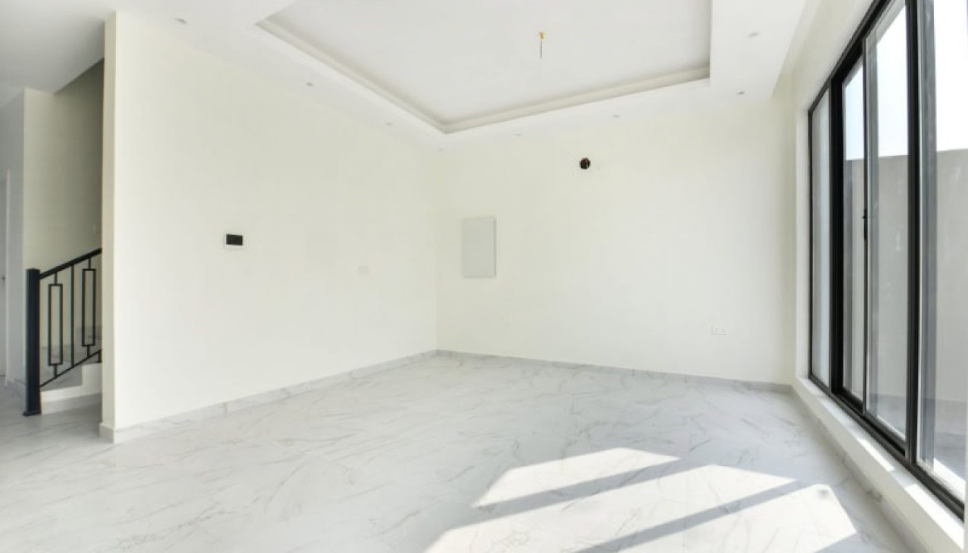 An empty room with white walls, marble floor, large windows on the right, and a partially visible staircase to the left.