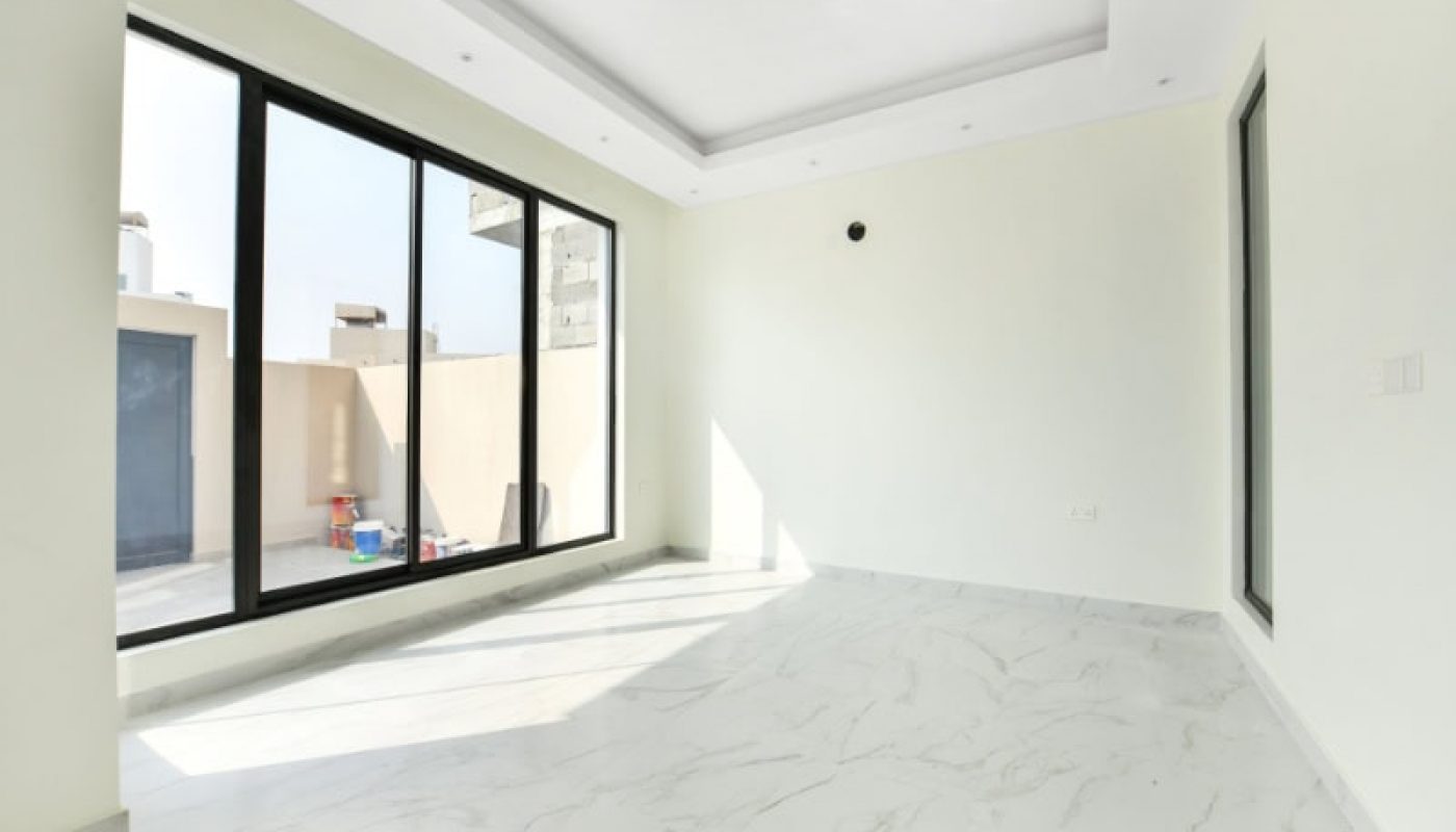 Empty room with large windows, white walls, and marble floor. Sunlight streams in, casting shadows on the floor. Boxes and toys visible outside the window on a balcony.