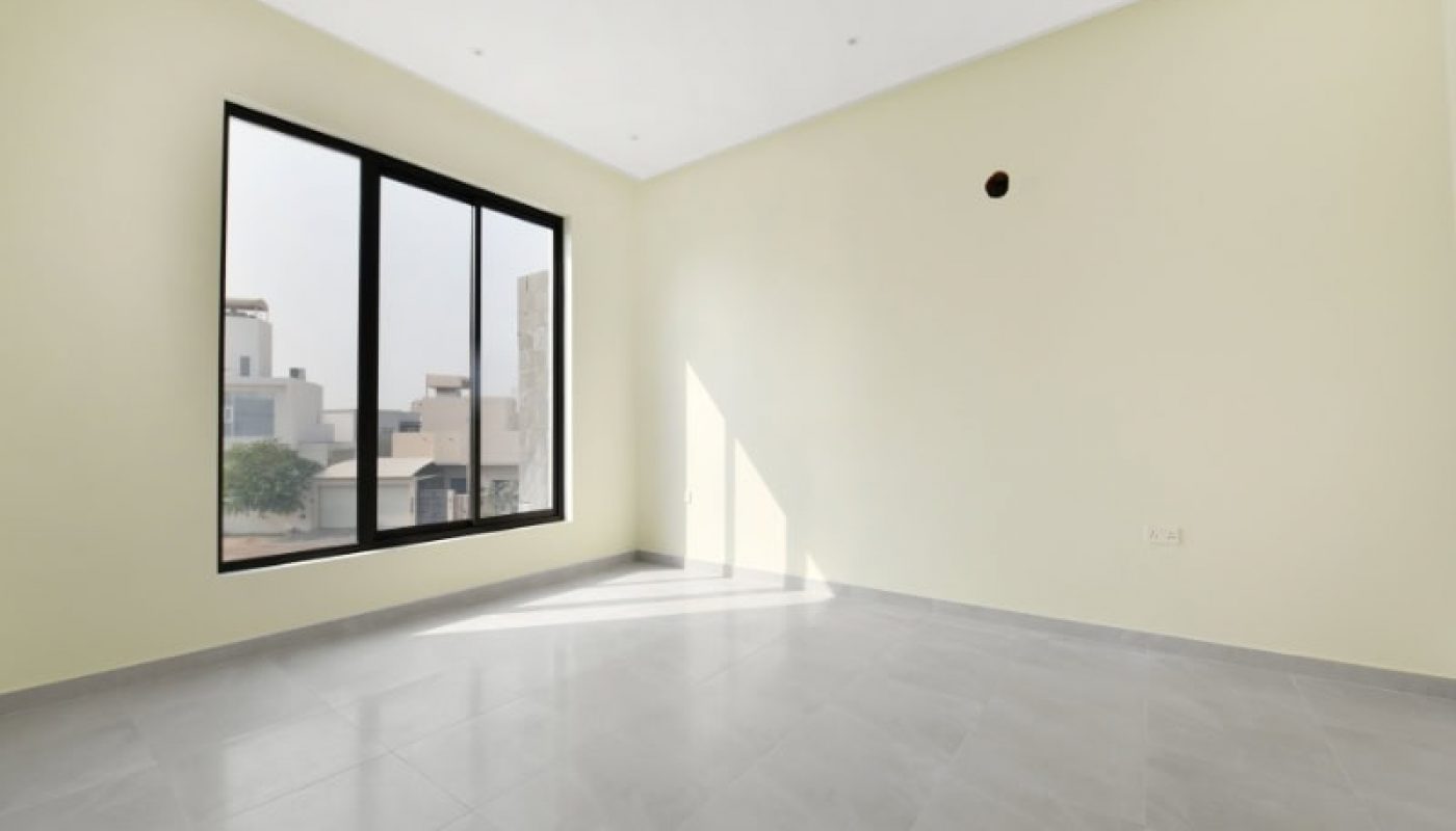 An empty room with a large window, pale yellow walls, and gray tiled floor. Light streams in from the window, casting shadows across the floor.