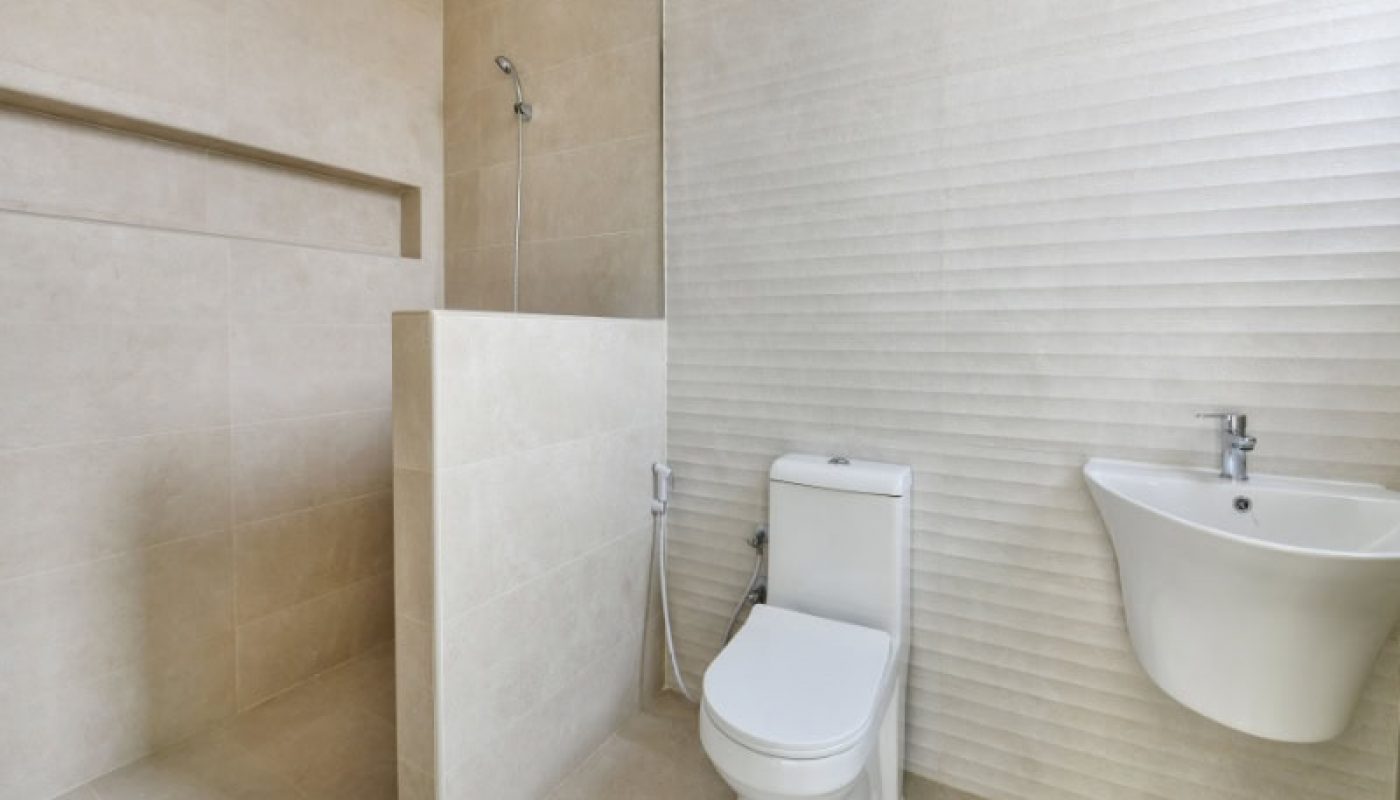 Modern bathroom with a toilet, wall-mounted sink, and open shower. Beige tiles cover the walls and floor.