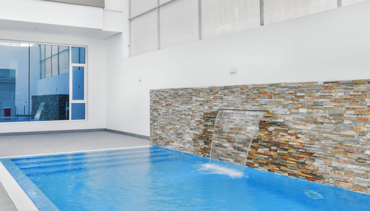 Indoor swimming pool with blue water, stone accent wall, and a waterfall feature in a bright, modern space with large windows and high ceilings.