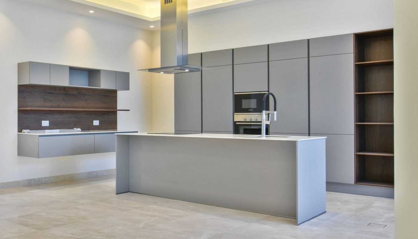 Modern kitchen with gray cabinets, built-in oven, central island with sink, open shelves, and tiled flooring. The space is empty and well-lit with recessed ceiling lights.