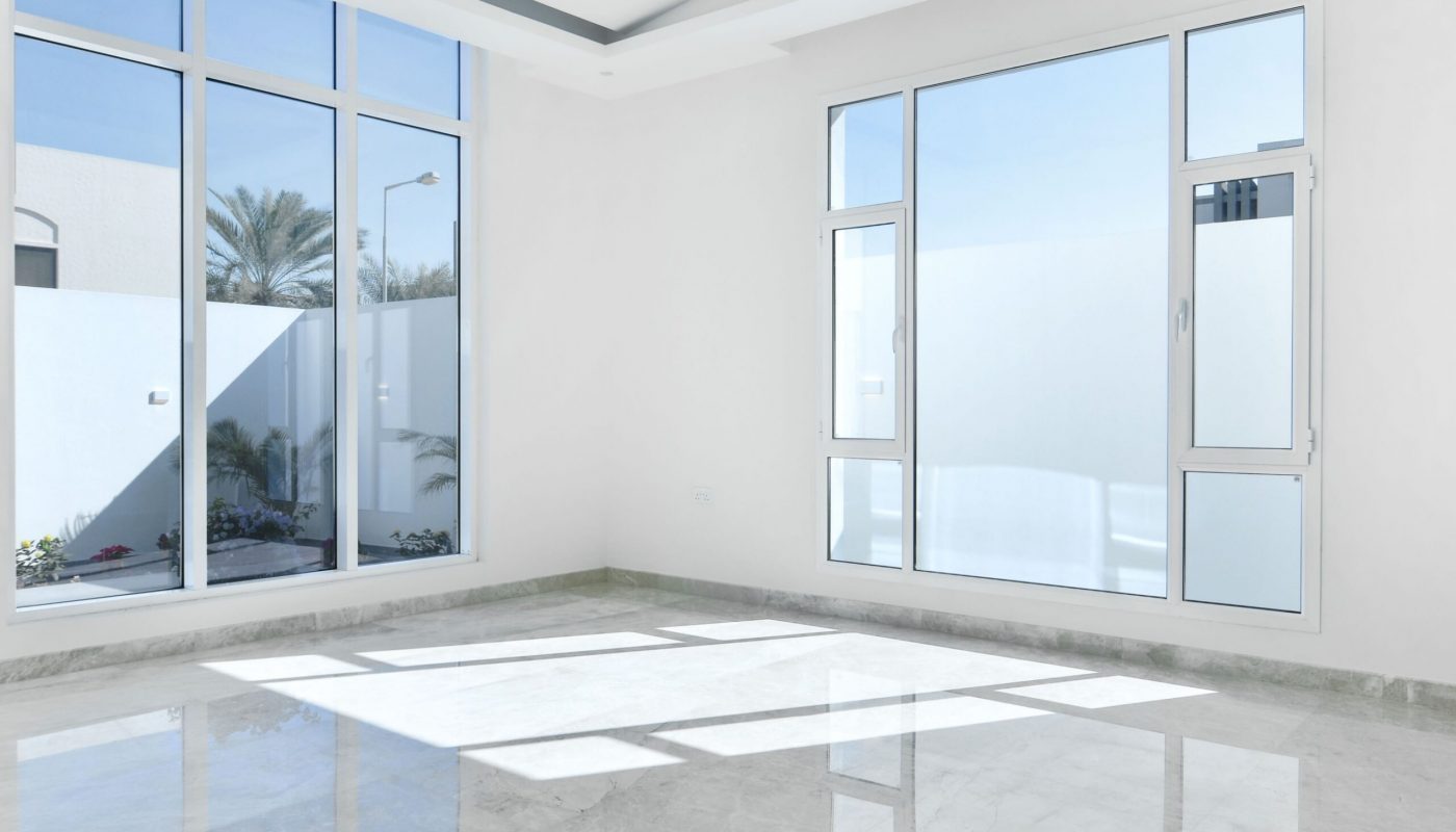 Empty modern room with large windows, white walls, and a shiny marble floor. Sunlight creates rectangular shadows on the ground.