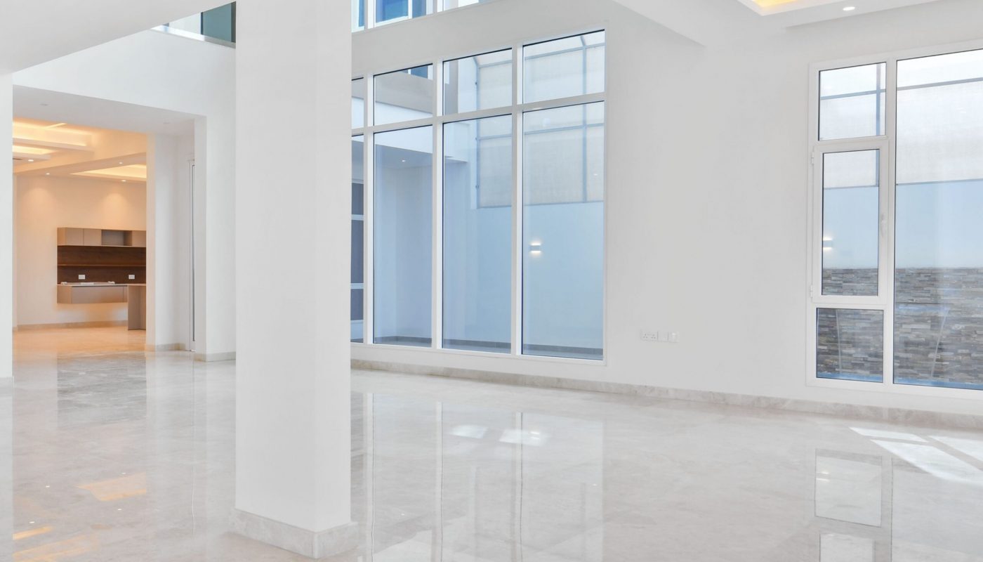 Spacious, modern interior with large windows, white walls, marble floors, and recessed ceiling lights. An open kitchen is visible in the background.