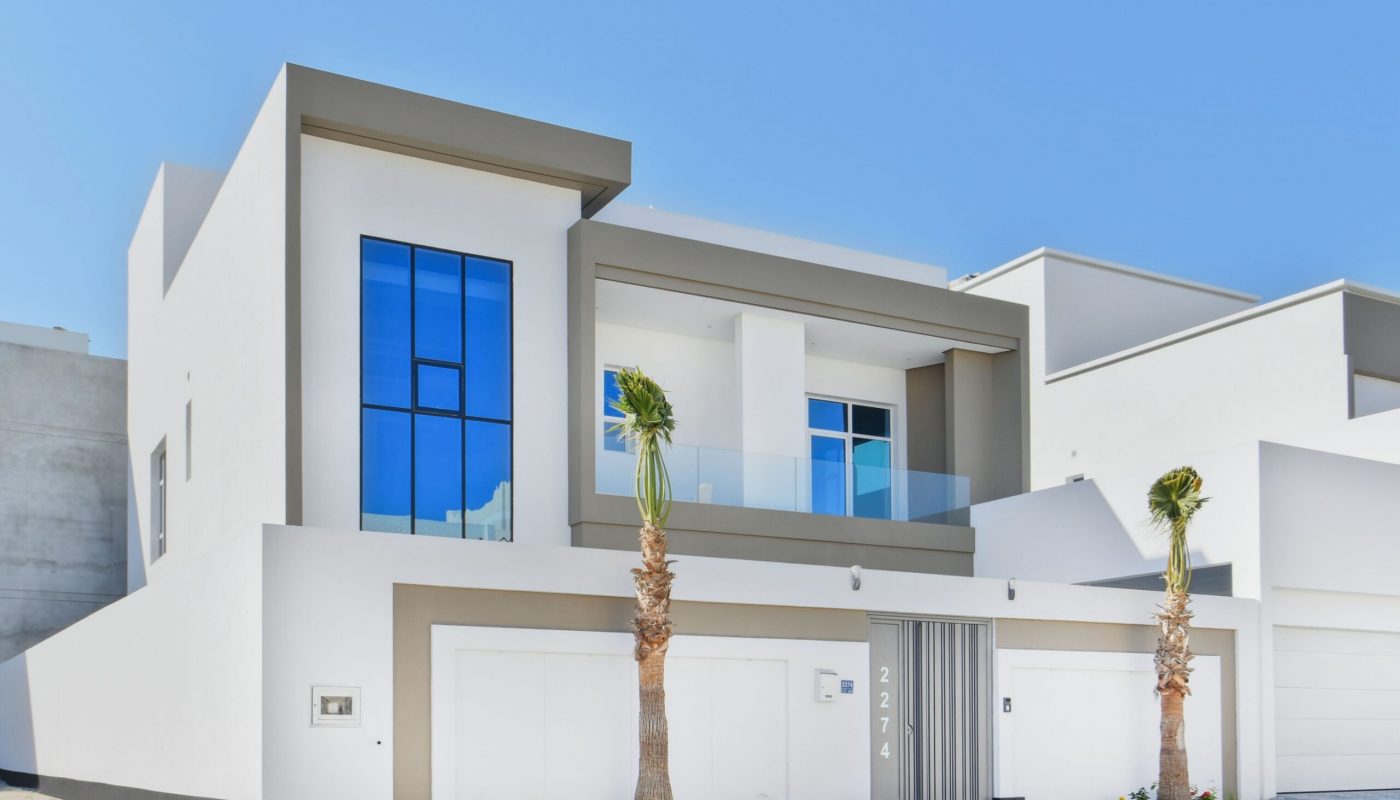 Modern two-story white house with large blue-tinted windows, a glass balcony, minimal landscaping, and two palm trees in front.
