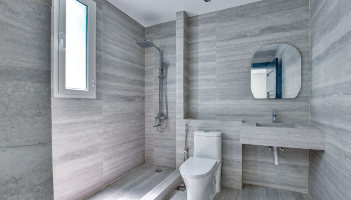 Modern bathroom with gray marble walls, a walk-in shower with a rainfall showerhead, a toilet, a round mirror, a sink, and a window allowing natural light.