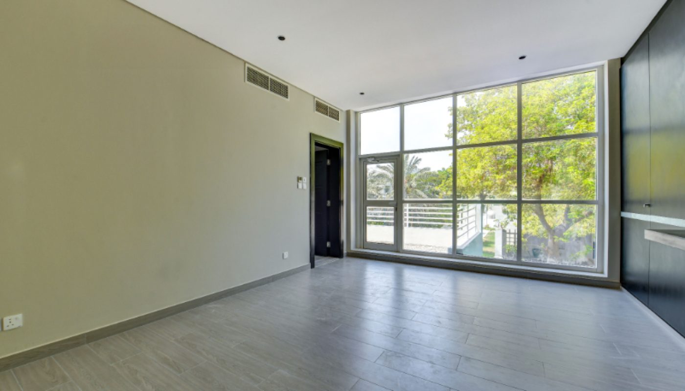 Empty room with light-colored tile flooring, large floor-to-ceiling window, neutral walls, and a door leading to another area. Greenery visible outside the window.