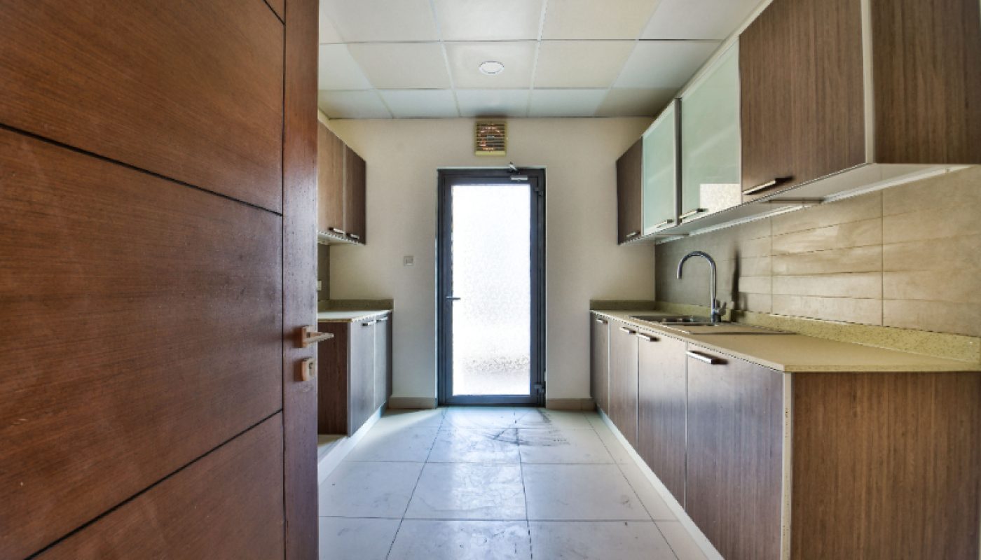 Modern galley kitchen with wooden cabinets, cream countertops, tiled floor, and a glass door at the far end letting in natural light.