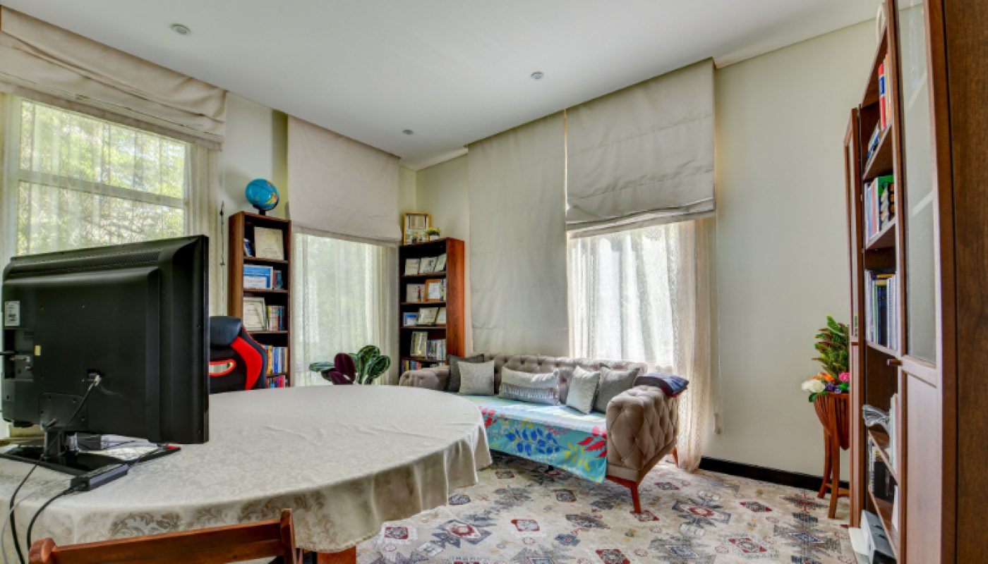 Home office with a round table, computer, wooden shelves with books, a daybed with cushions, large windows with shades, and a patterned rug.