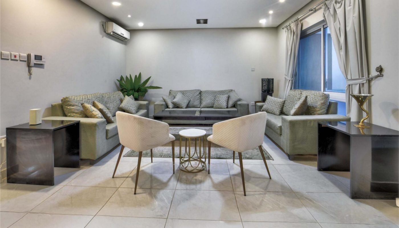 Modern living room with tiled floor, gray sectional sofa, two white chairs, a round coffee table, side tables, potted plant, and window with curtains. Ceiling lights are on.