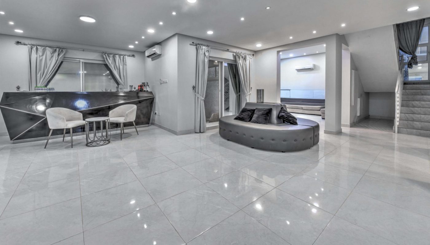 Spacious modern living room with glossy white tiled floor, a round gray sofa, a bar area with two chairs, and gray curtains on windows and entryways.