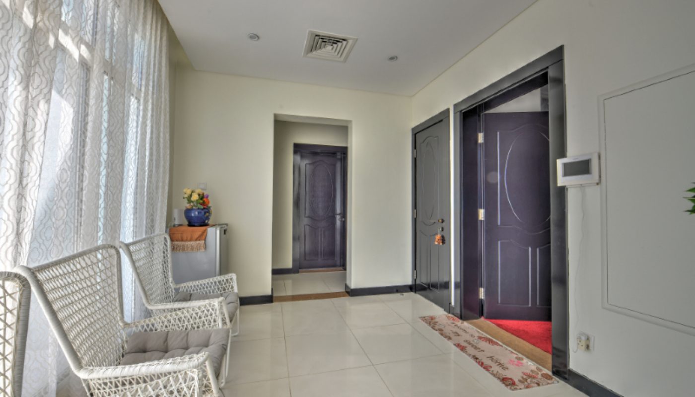 A hallway with white tile flooring, wicker chairs by windowed curtains, two dark doors, and a wall-mounted control panel.