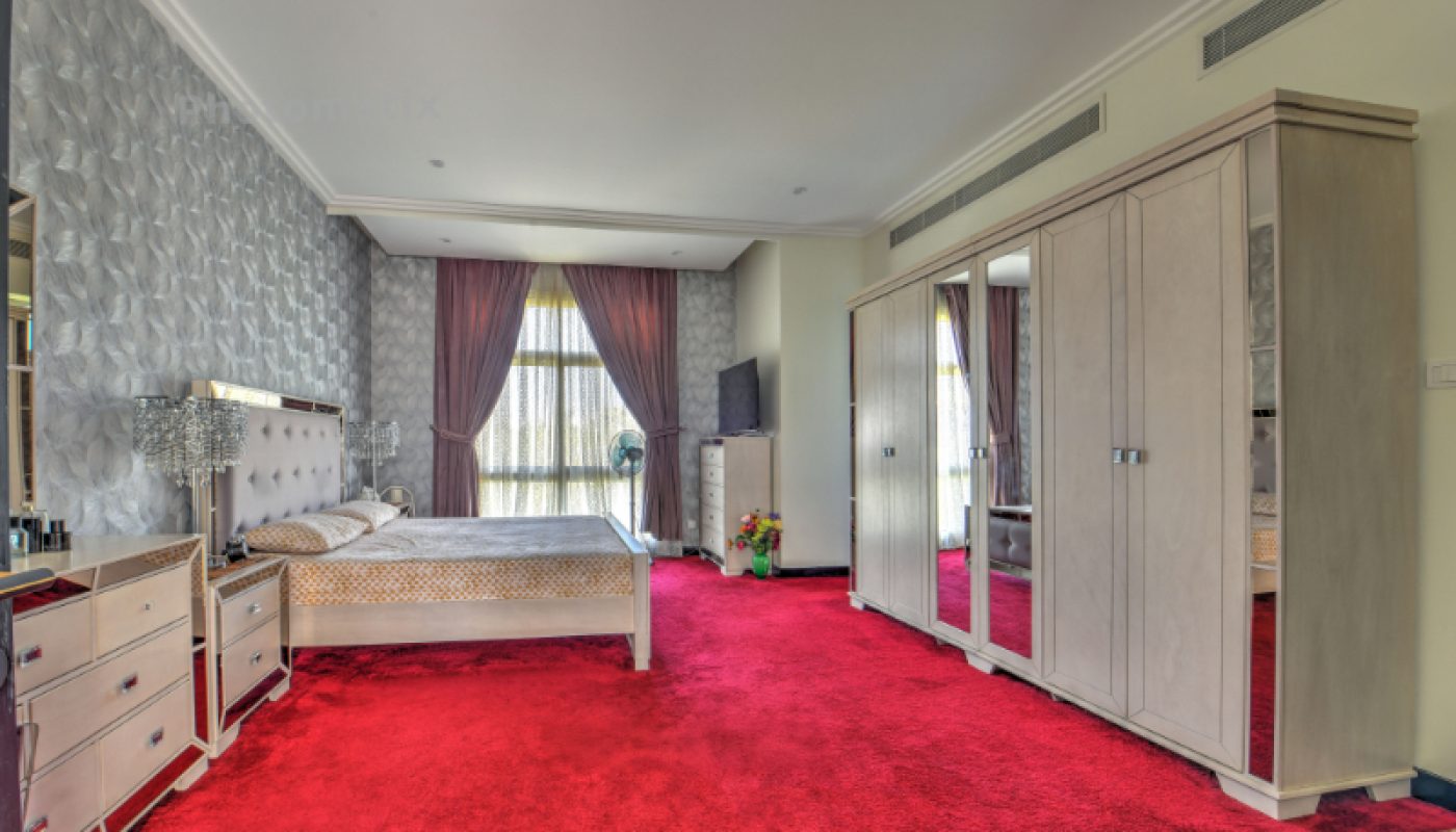 Spacious bedroom with red carpet, large bed, multiple wardrobes, dresser, and a window with sheer and dark curtains letting in natural light.