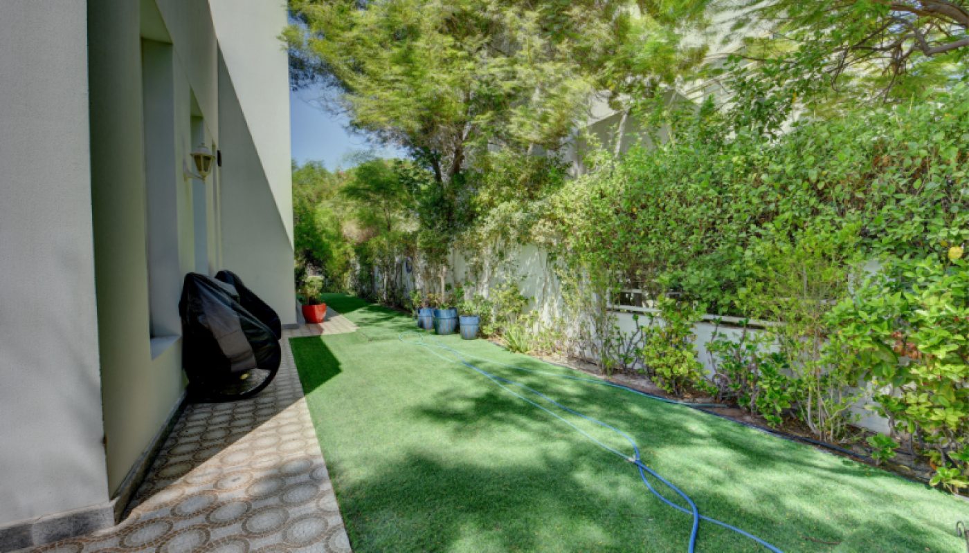 A narrow backyard with artificial grass, green shrubs, potted plants, a blue hose on the ground, and a chair next to the house wall.