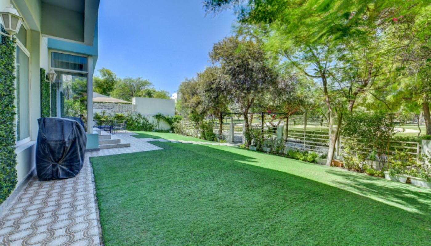A backyard with artificial grass, bordered by trees and a fence, features a covered grill and outdoor seating area near a house with large windows.