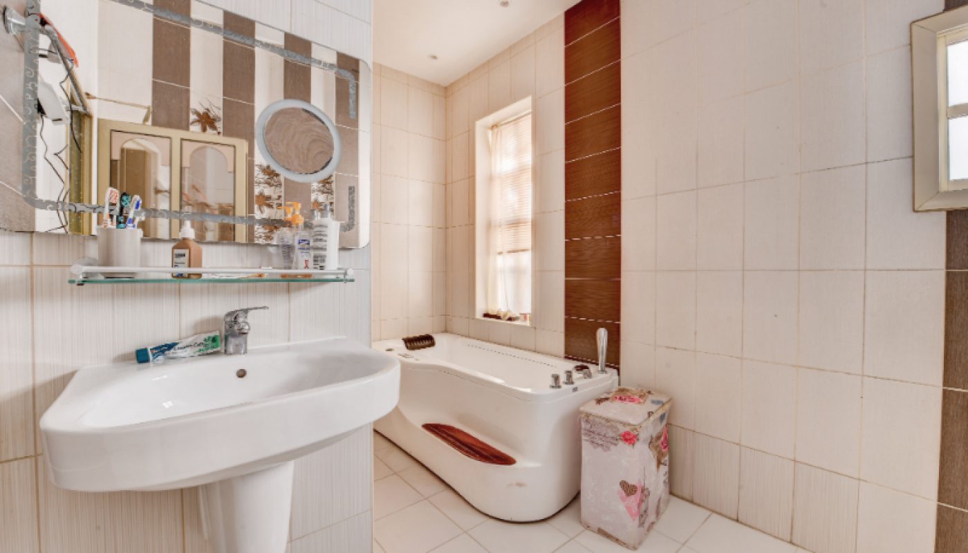 Modern bathroom with a white sink, mirror, glass shelf with toiletries, a bathtub, a window with blinds, and a covered laundry basket.