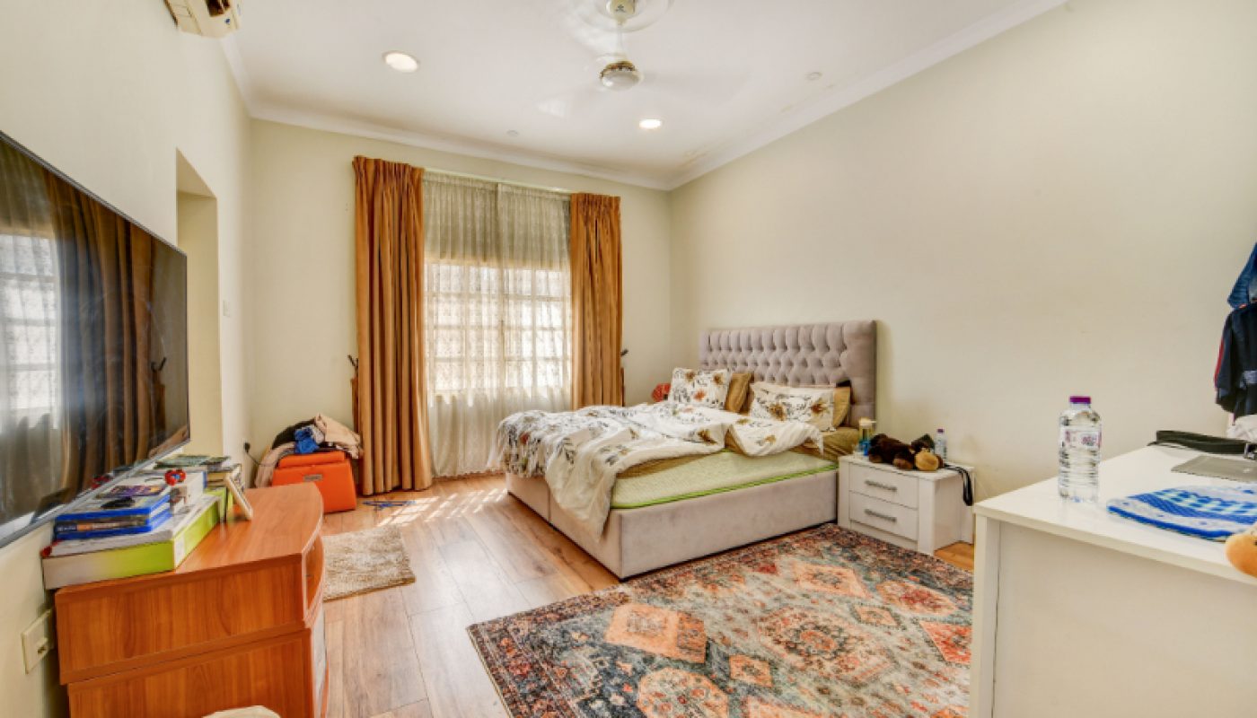 A well-lit bedroom with a double bed, large rug, wall-mounted TV, wooden furniture, and curtains covering a window letting in natural light.