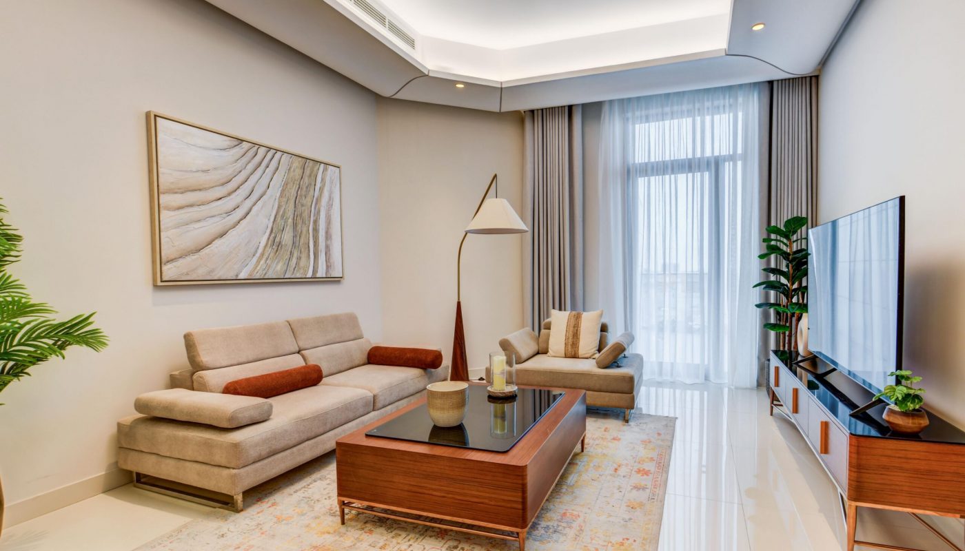Modern living room with beige sofas, a wooden coffee table, floor lamp, large TV, potted plants, abstract wall art, and floor-to-ceiling curtains covering a tall window.