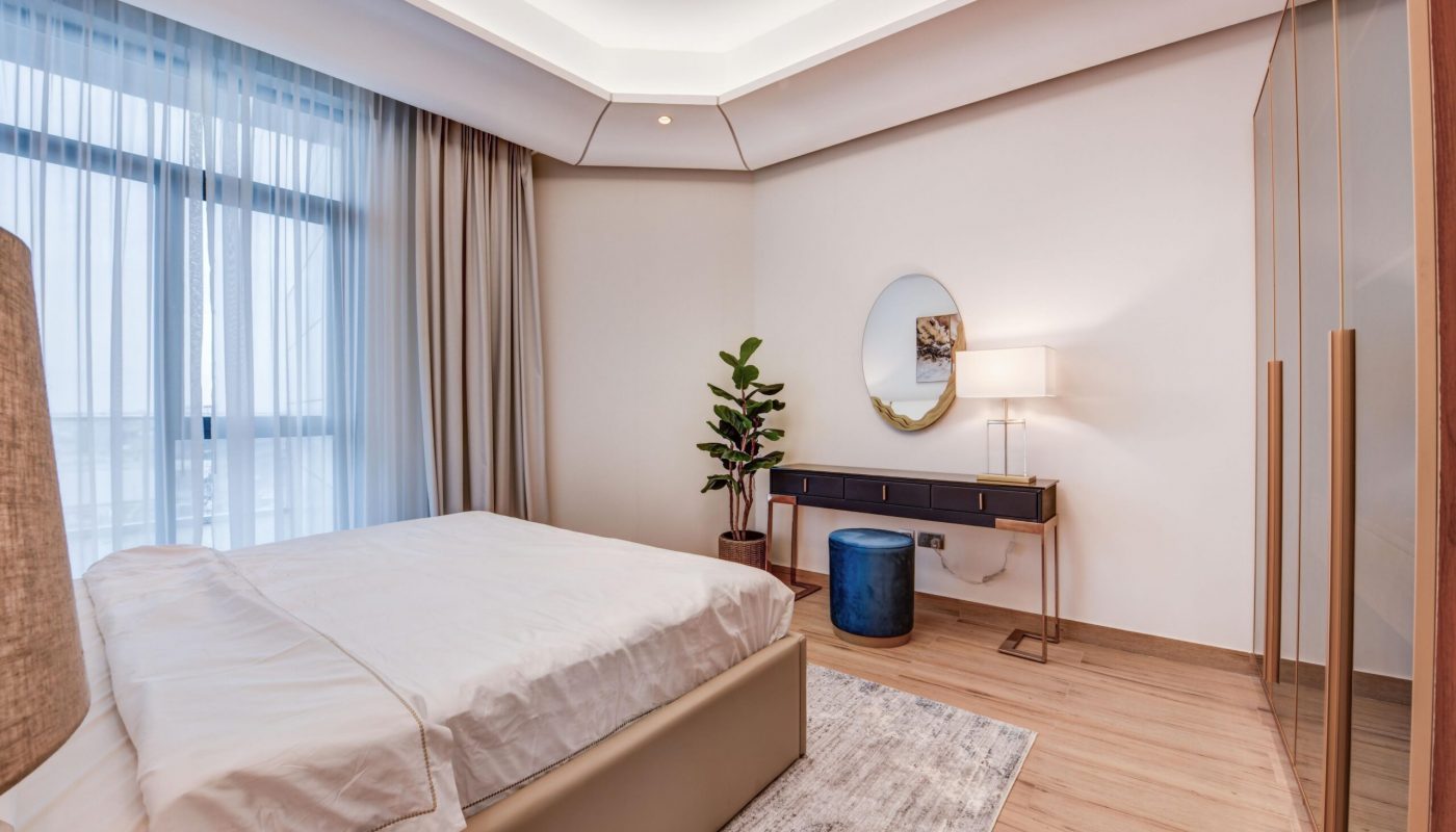 Modern bedroom with a large bed, wooden floors, a dressing table with a round mirror, a lamp, a potted plant, and floor-to-ceiling curtained windows.