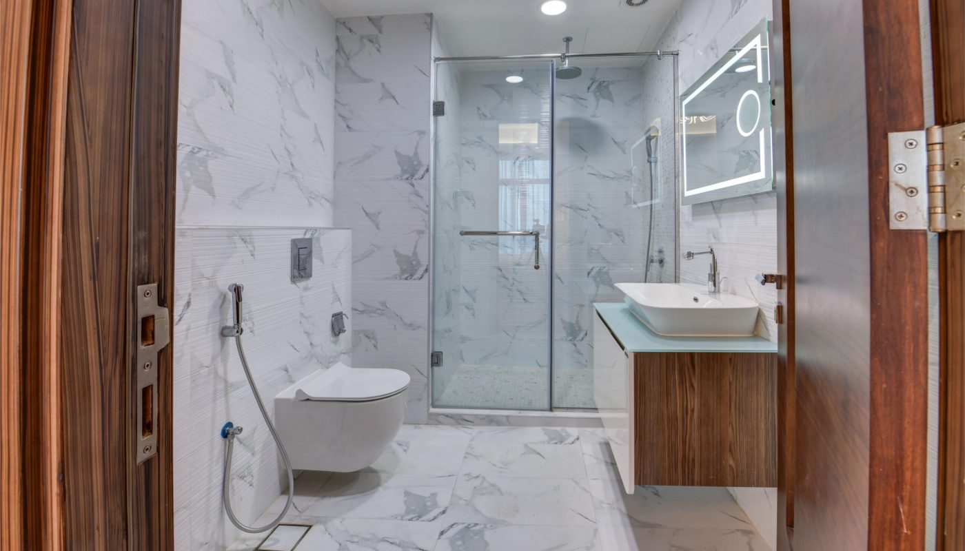 Modern bathroom with white marble walls and floor, wall-mounted toilet, glass shower enclosure, floating vanity with vessel sink, and large backlit mirror.