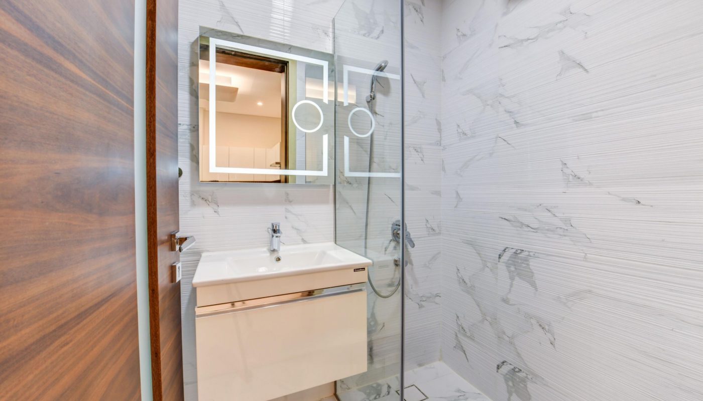 Modern bathroom with a wall-mounted sink, illuminated mirror, glass shower enclosure, and white marble-patterned tiles.