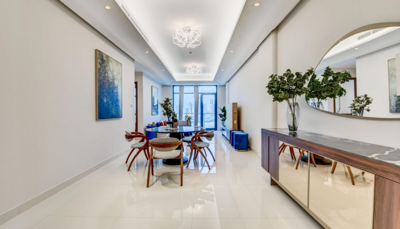Modern dining room with white floors, wooden chairs around a table, a sideboard with a round mirror, plants, and abstract art on the walls. Large windows let in natural light.