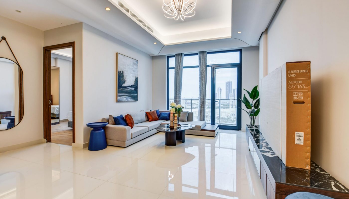 Modern living room with a sectional sofa, coffee table, large windows with city view, wall art, and a boxed Samsung TV on a marble console. Doorways lead to adjacent rooms.