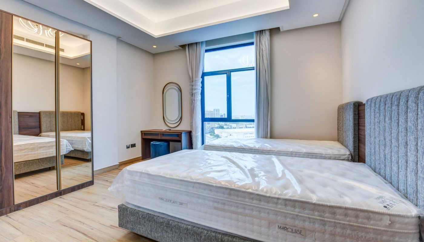 Modern bedroom with two single beds, a mirrored wardrobe, a small desk with a mirror, large window with city view, and wooden flooring.